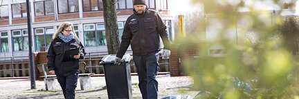 Zwei Mitarbeitende des Kontrolldienstes prüfen illegale Müllablagerungen auf der Straße. Eine Person zieht eine Tonne hinter sich her. | © Die Bremer Stadtreinigung