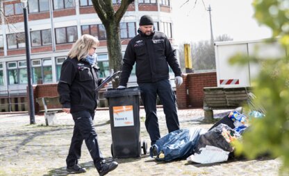 Zwei Mitarbeitende des Kontrolldienstes blicken auf einen Haufen illegaler Müllablagerungen. Die linke Person hat ein Klemmbrett in der Hand und die rechte Person hat eine Mülltonne zwischen ihnen abgestellt. | © Die Bremer Stadtreinigung