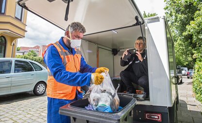 Im Wagen des Kontrolldienstes sitzt eine Frau und fotografiert mit dem Handy. Vor dem geöffneten Kofferraum zieht ihr Kollege eine Mülltüte aus einer Restmülltonne. | © Die Bremer Stadtreinigung