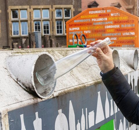 Eine Person wirft eine durchsichtige Glasflasche in die Öffnung eines Containers. Auf dem Container steht Weißglas. | © Die Bremer Stadtreinigung