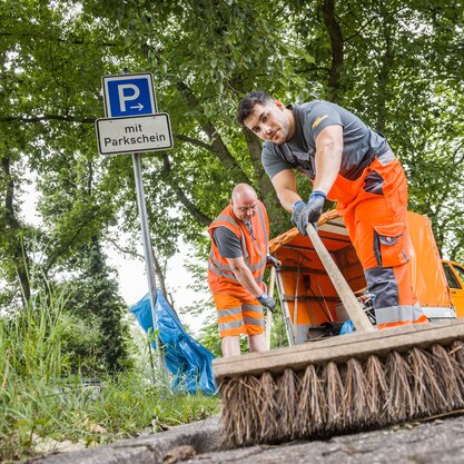 Zwei Mitarbeitende von DBS fegen mit einem Besen die Straße. In der Bildmitte lehnt ein Blauer Müllsack an einem Straßenschild, das auf eine Parkfläche hinweist. | © Die Bremer Stadtreinigung